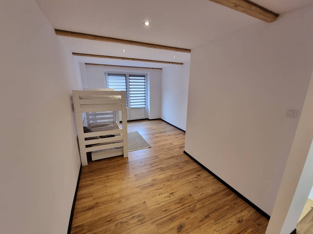 Pokój z drewnianą podłogą, białymi ścianami, drewnianymi belkami stropowymi i białym łóżkiem piętrowym w rogu.