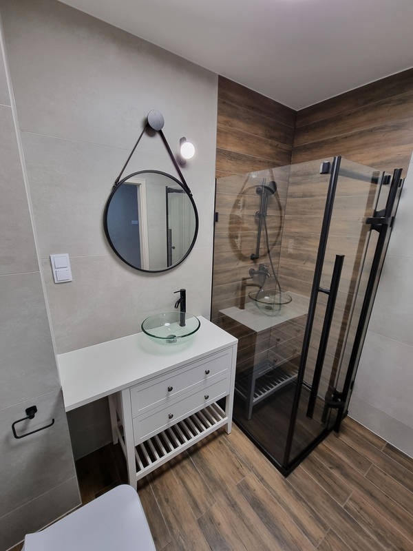 Nowoczesna łazienka z kabiną prysznicową w czarnej ramie, umywalką nablatową ze szkła na białej szafce z szufladami i okrągłym lustrem zawieszonym na skórzanym pasku.