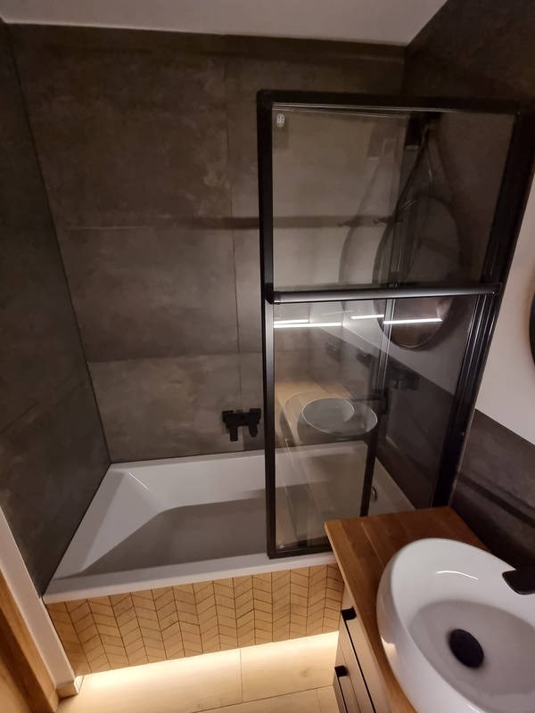 Nowoczesna łazienka z wanną obudowaną drewnopodobnymi płytkami ułożonymi w jodełkę, czarna armatura i szklany parawan nawannowy z czarną ramą, umywalka nablatowa na drewnianej szafce.