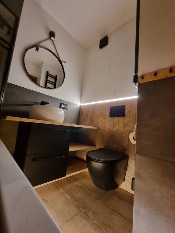 Nowoczesna łazienka z wiszącą czarną toaletą, umywalką nablatową na drewnianym blacie i okrągłym lustrem zawieszonym na sznurze, podświetlona listwą LED.