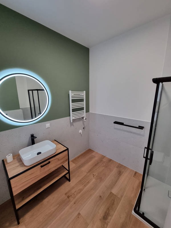 Nowoczesna łazienka z umywalką nablatową na drewnianej szafce z czarnymi elementami, okrągłe lustro z podświetleniem LED, grzejnik drabinkowy i kabina prysznicowa z czarnymi profilami.