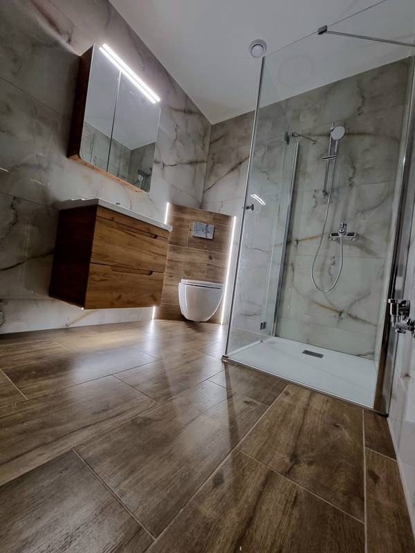 Nowoczesna łazienka z drewnopodobnymi płytkami na podłodze, marmurowymi płytkami na ścianach, podwieszaną szafką z umywalką, toaletą w zabudowie i szklanym prysznicem typu walk-in.