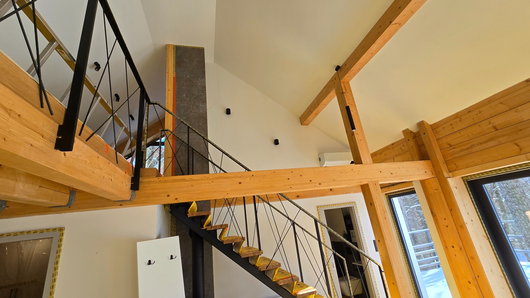 Wnętrze domu w trakcie wykańczania, widoczne drewniane belki stropowe, czarne schody z zabezpieczonymi stopniami i metalową balustradą, ściany przygotowane do malowania, okna z widokiem na zimowy...