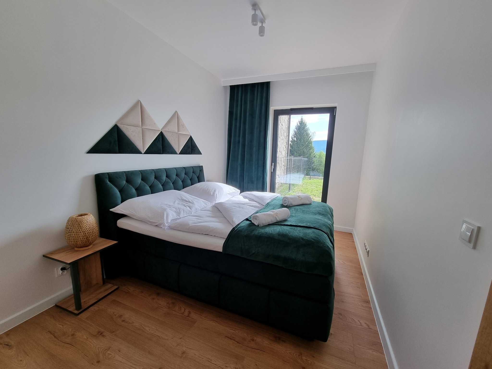 Nowoczesna sypialnia z zielonym łóżkiem z pikowanym zagłówkiem, drewnianą podłogą i dekoracją ścienną w kształcie gór, widok na zewnątrz przez okno balkonowe.