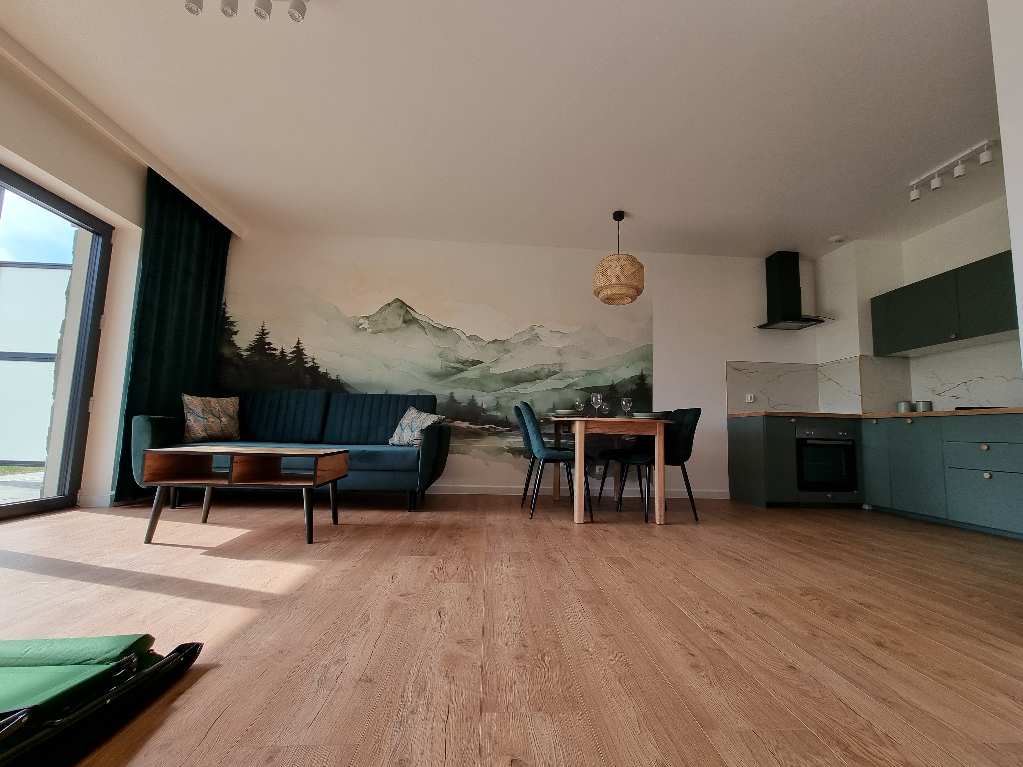 Wnętrze salonu z aneksem kuchennym, ściana z motywem górskim, drewniana podłoga, zielona sofa i krzesła, widok z niskiego kąta, częściowo złożone łóżko turystyczne na pierwszym planie.