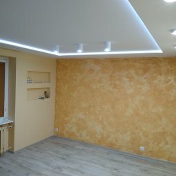 Grubud - Wykończone wnętrze pokoju z dekoracyjną ścianą w odcieniach brązu i beżu, podświetlanym sufitem z oświetleniem LED, grzejnikiem żeberkowym przy oknie i panelami podłogowymi imitującymi drewno.