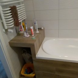 Grubud - Fragment łazienki z wanną, grzejnikiem drabinkowym z brązowym ręcznikiem, szafką z akcesoriami do higieny jamy ustnej i żółtym koszem na śmieci pod szafką.