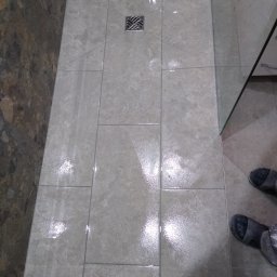 Grubud - Wykończony brodzik z odpływem liniowym, wyłożony beżowymi płytkami imitującymi kamień, częściowo osłonięty szklaną ścianką kabiny prysznicowej. Na podłodze widoczne ślady wody i robocze obuwie.