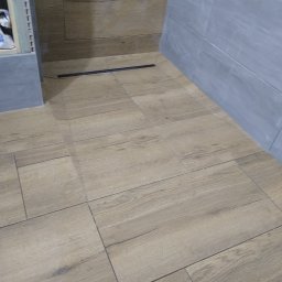 Grubud - Ułożona podłoga z płytek imitujących drewno w odcieniach beżu, z widocznym odpływem liniowym i fragmentem szarej ściany wyłożonej płytkami.