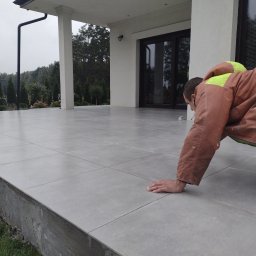 Grubud - Układanie szarych płytek na tarasie domu jednorodzinnego; pracownik w kombinezonie z elementami odblaskowymi sprawdza równość powierzchni.