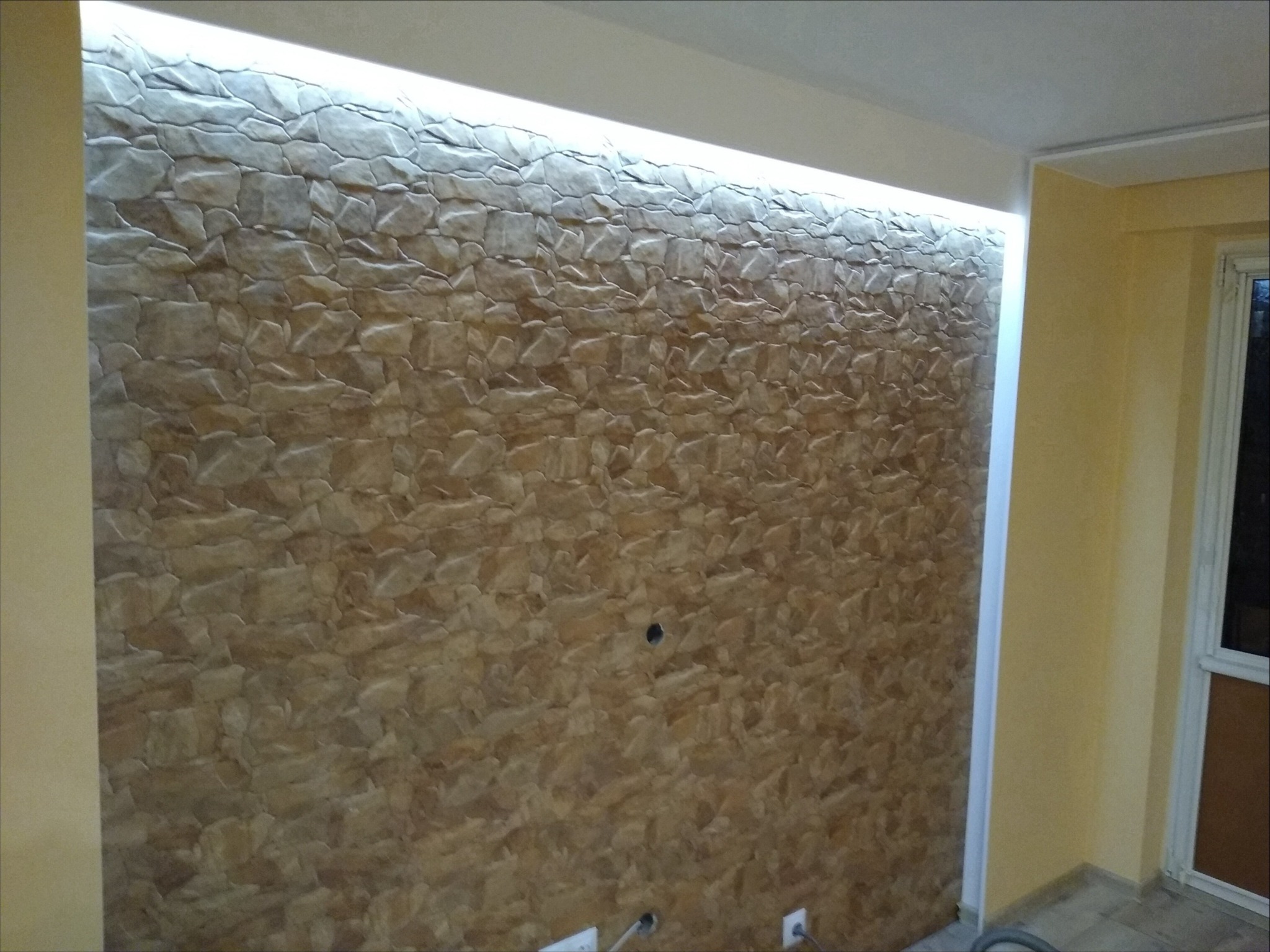 Ściana wykończona dekoracyjną okładziną imitującą kamień z podświetleniem LED w górnej części, widoczne gniazdka elektryczne w dolnej części ściany.