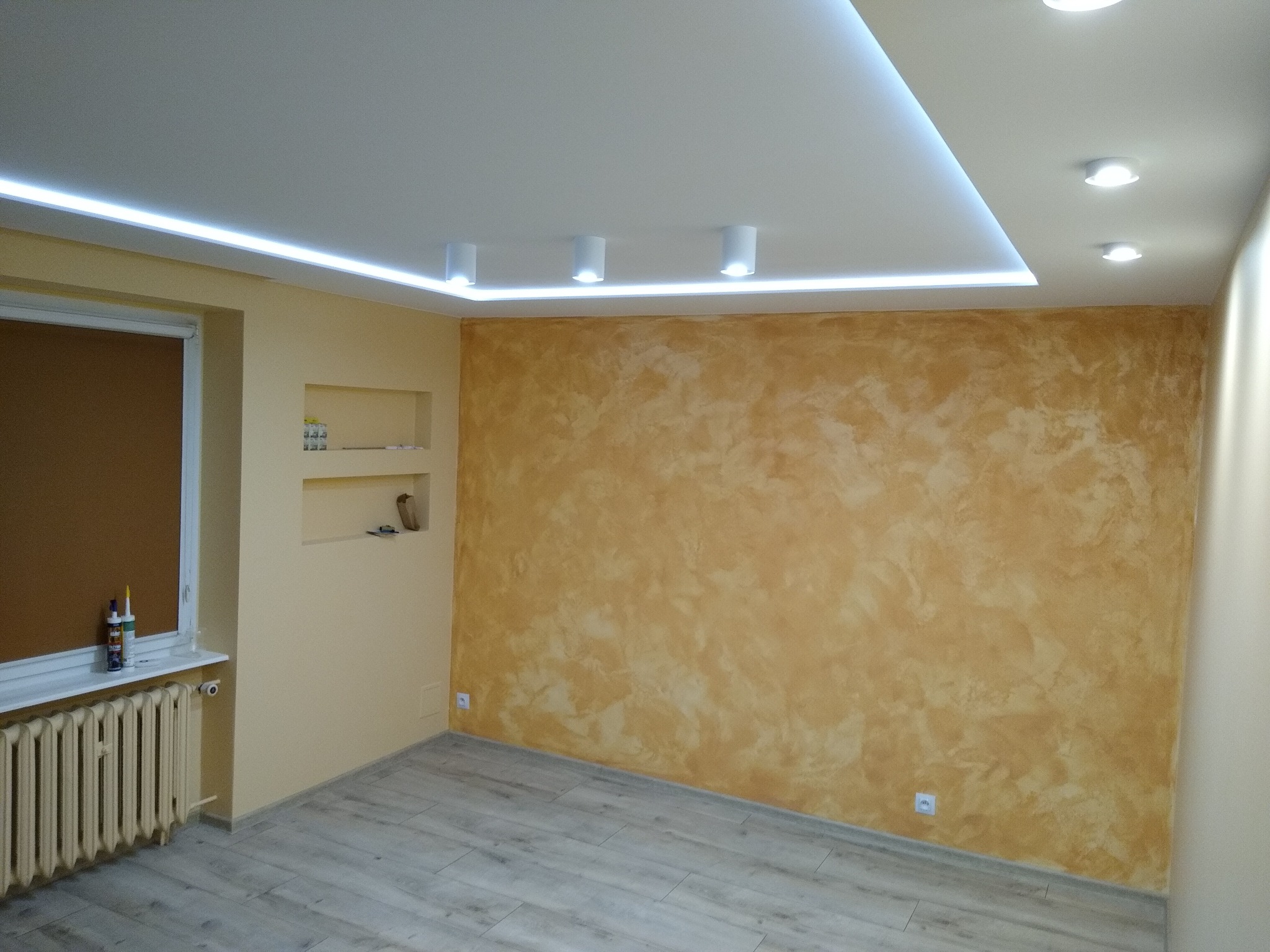 Wykończone wnętrze pokoju z dekoracyjną ścianą w odcieniach brązu i beżu, podświetlanym sufitem z oświetleniem LED, grzejnikiem żeberkowym przy oknie i panelami podłogowymi imitującymi drewno.