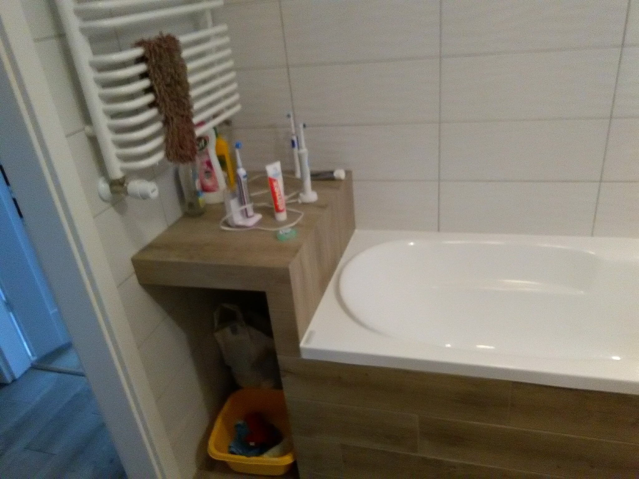 Fragment łazienki z wanną, grzejnikiem drabinkowym z brązowym ręcznikiem, szafką z akcesoriami do higieny jamy ustnej i żółtym koszem na śmieci pod szafką.