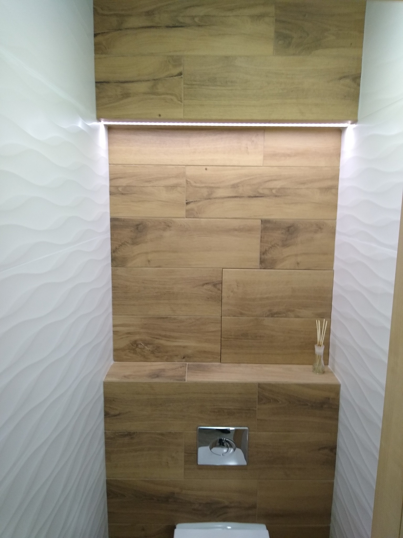 Nowoczesna toaleta z drewnopodobnymi płytkami na ścianie akcentującej, podświetleniem LED i dekoracyjnymi białymi płytkami 3D na sąsiedniej ścianie.