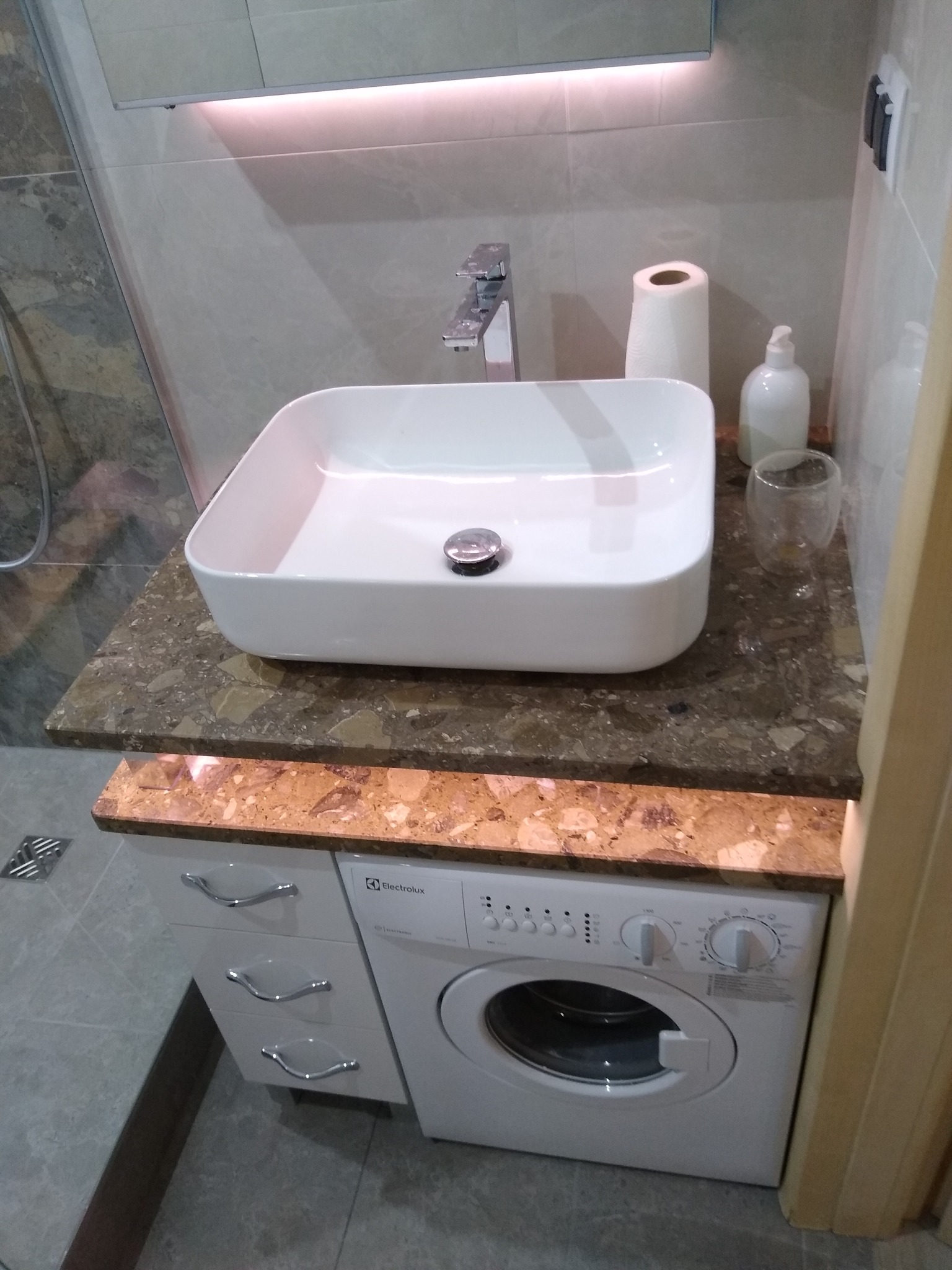 Nowoczesna łazienka z prostokątną umywalką nablatową na blacie z brązowego lastryko, podświetlonym od dołu, oraz pralką Electrolux w zabudowie szafkowej z trzema szufladami.
