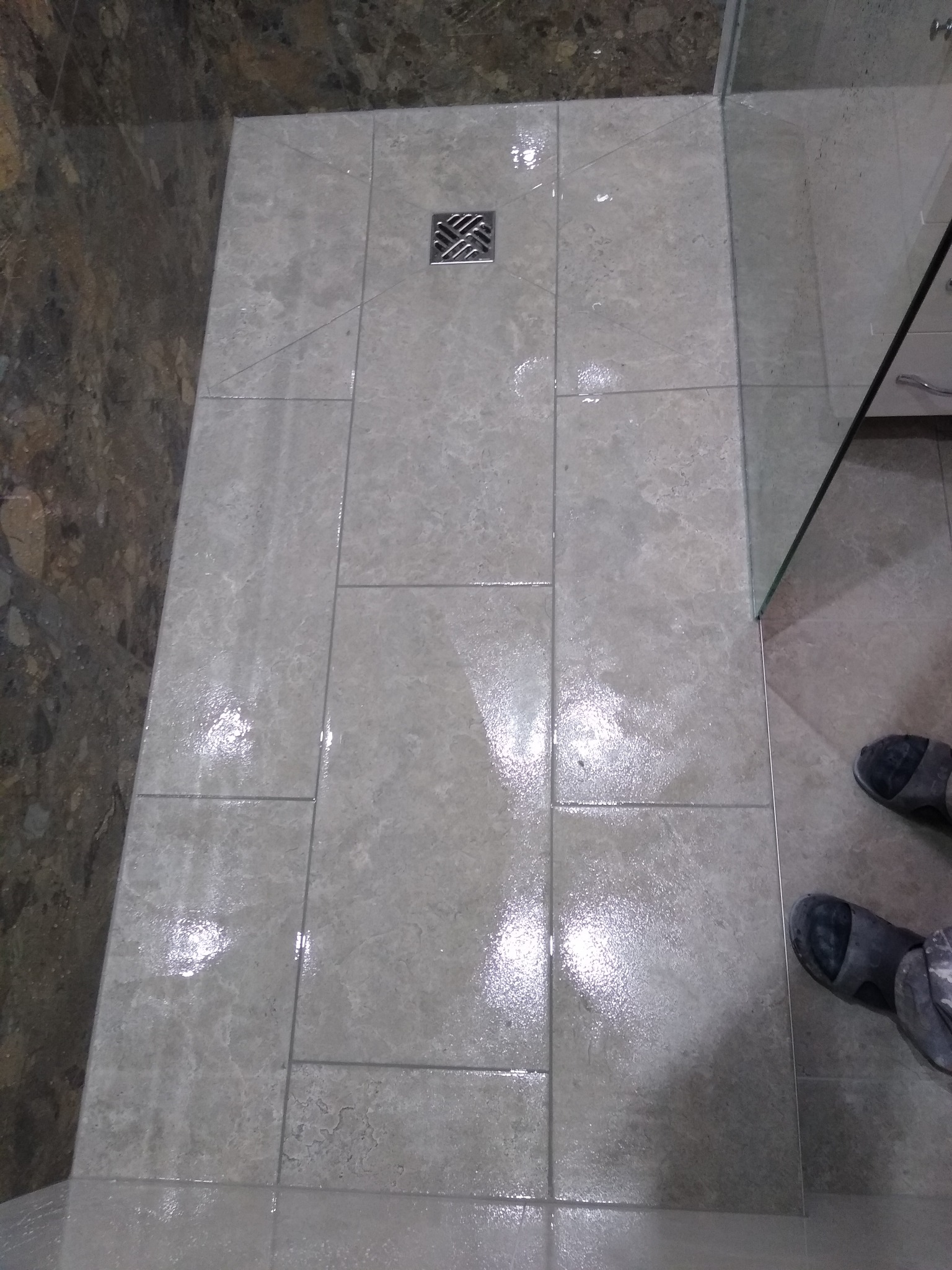 Wykończony brodzik z odpływem liniowym, wyłożony beżowymi płytkami imitującymi kamień, częściowo osłonięty szklaną ścianką kabiny prysznicowej. Na podłodze widoczne ślady wody i robocze obuwie.