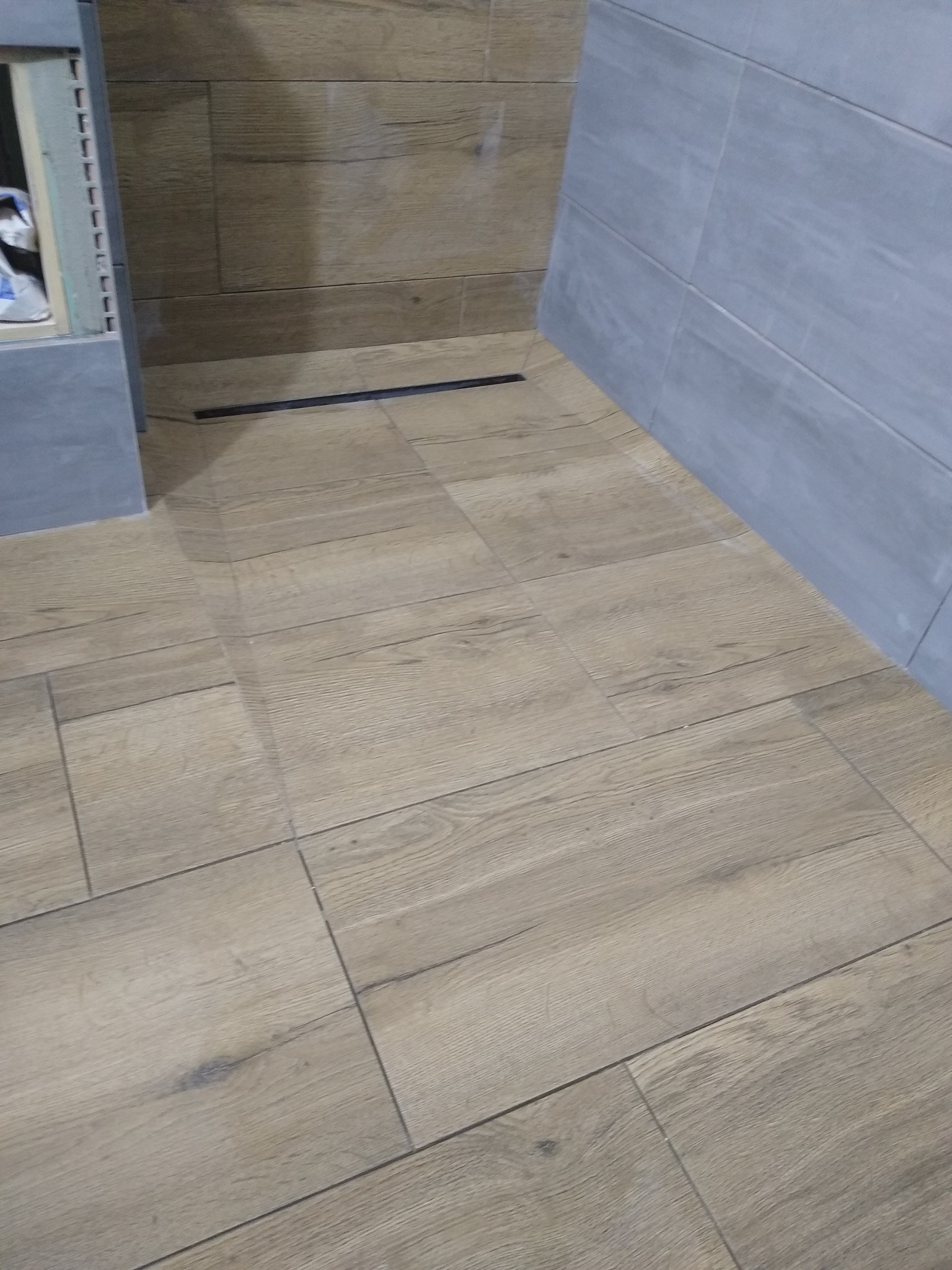 Ułożona podłoga z płytek imitujących drewno w odcieniach beżu, z widocznym odpływem liniowym i fragmentem szarej ściany wyłożonej płytkami.