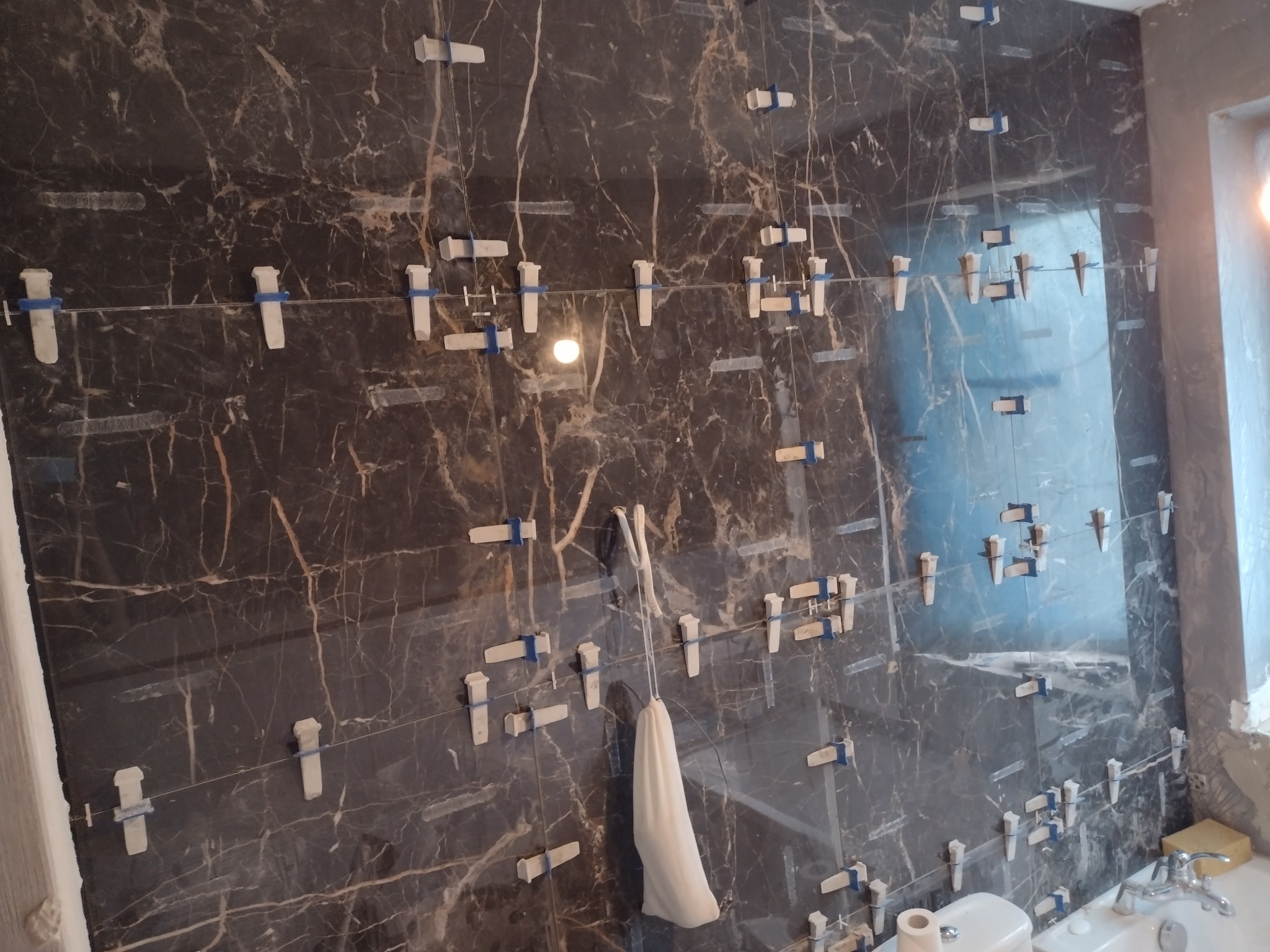 Ściana łazienki wyłożona ciemnymi płytkami imitującymi marmur, z widocznymi krzyżykami dystansowymi i sznurkiem poziomującym, sugerującymi etap układania płytek.