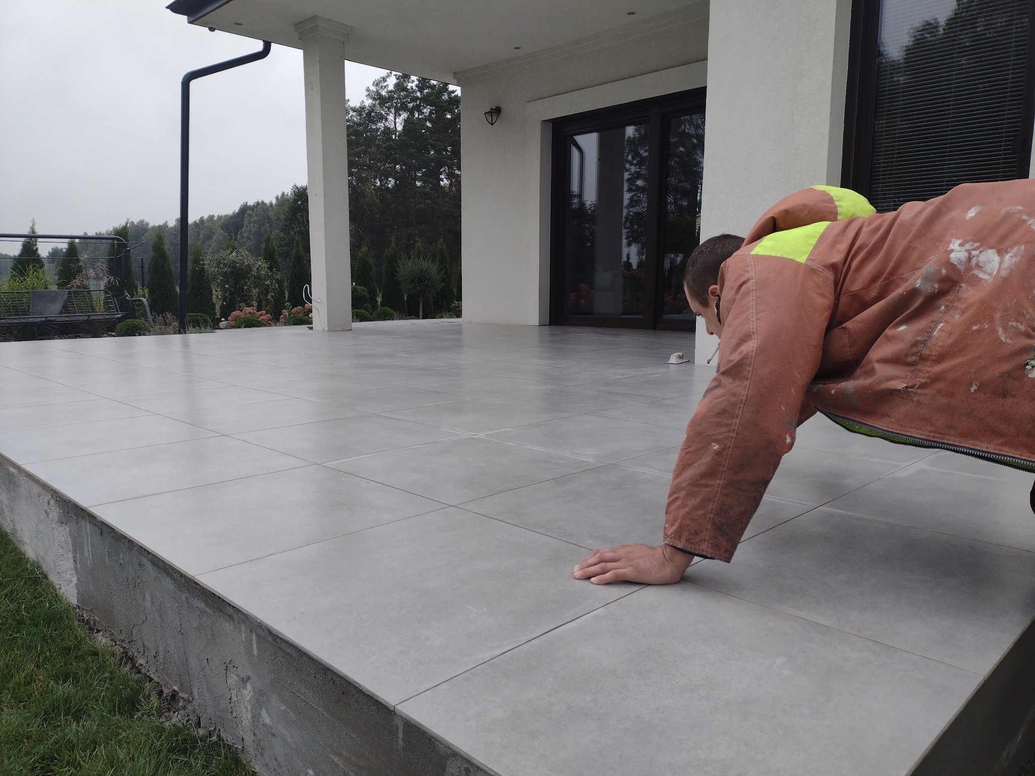 Układanie szarych płytek na tarasie domu jednorodzinnego; pracownik w kombinezonie z elementami odblaskowymi sprawdza równość powierzchni.