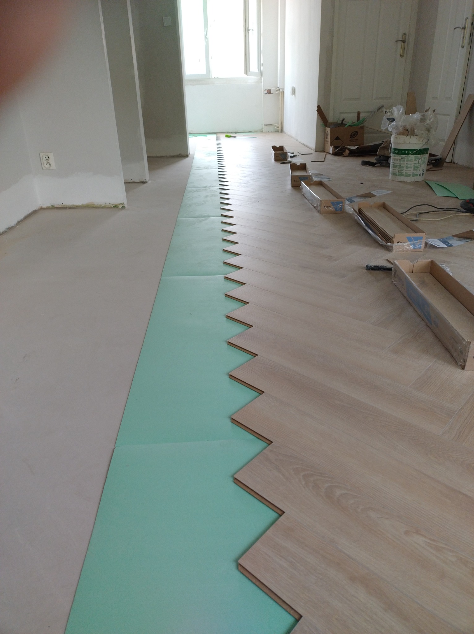 Układanie parkietu w jodełkę na zielonej piance izolacyjnej, widoczne puste kartony po panelach i wiadro z klejem.