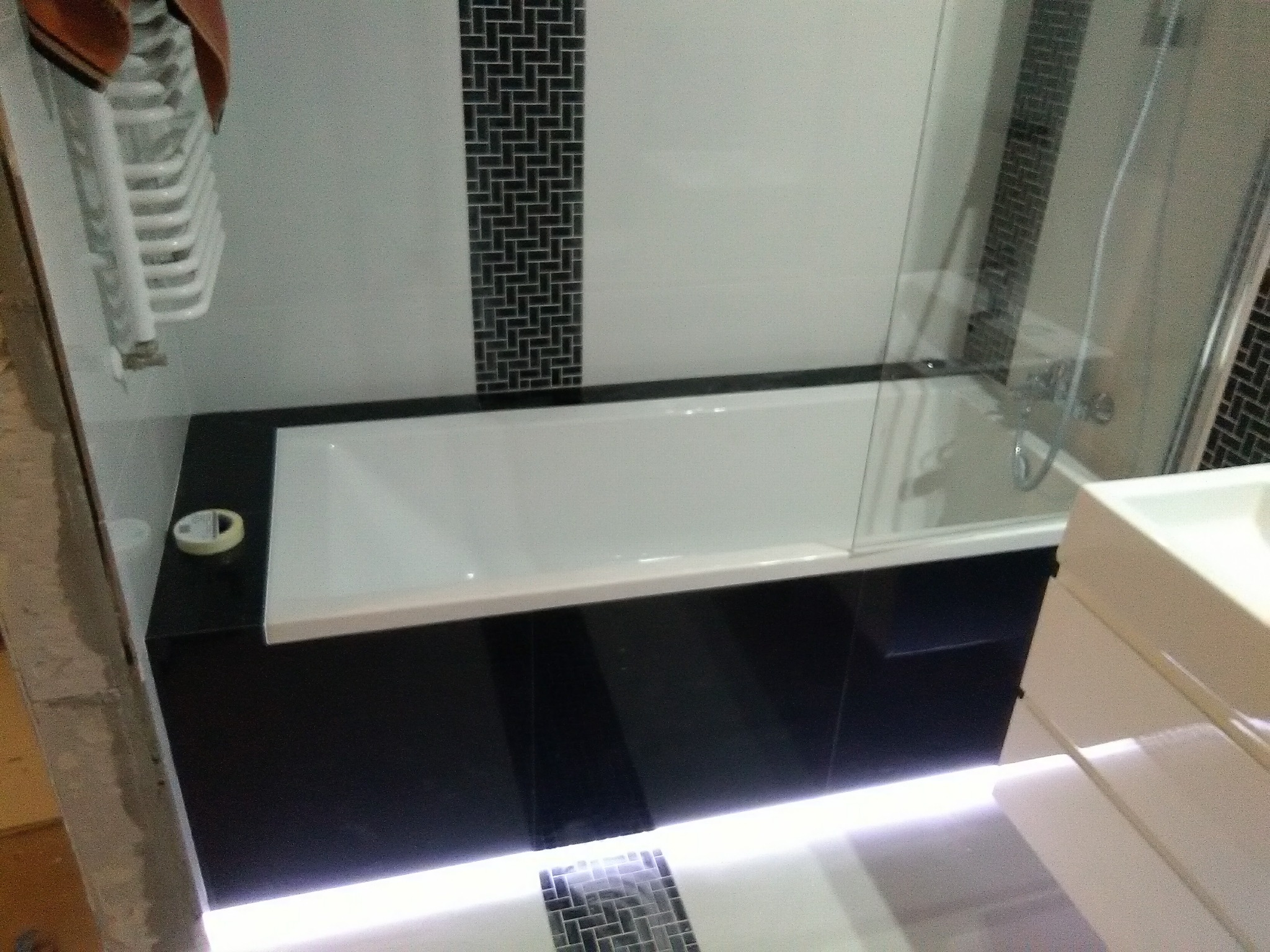 Nowoczesna łazienka z białą wanną obudowaną czarnymi płytkami, oświetleniem LED pod wanną i szklanym panelem prysznicowym, fragment grzejnika drabinkowego z ręcznikiem w rogu.