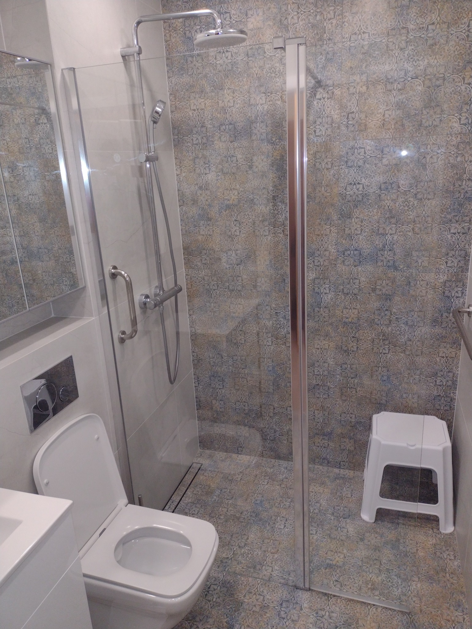 Wykończona łazienka z kabiną prysznicową ze szklanymi drzwiami, toaletą podwieszaną, wzorzystymi płytkami ściennymi i podłogowymi oraz małym białym stołkiem.