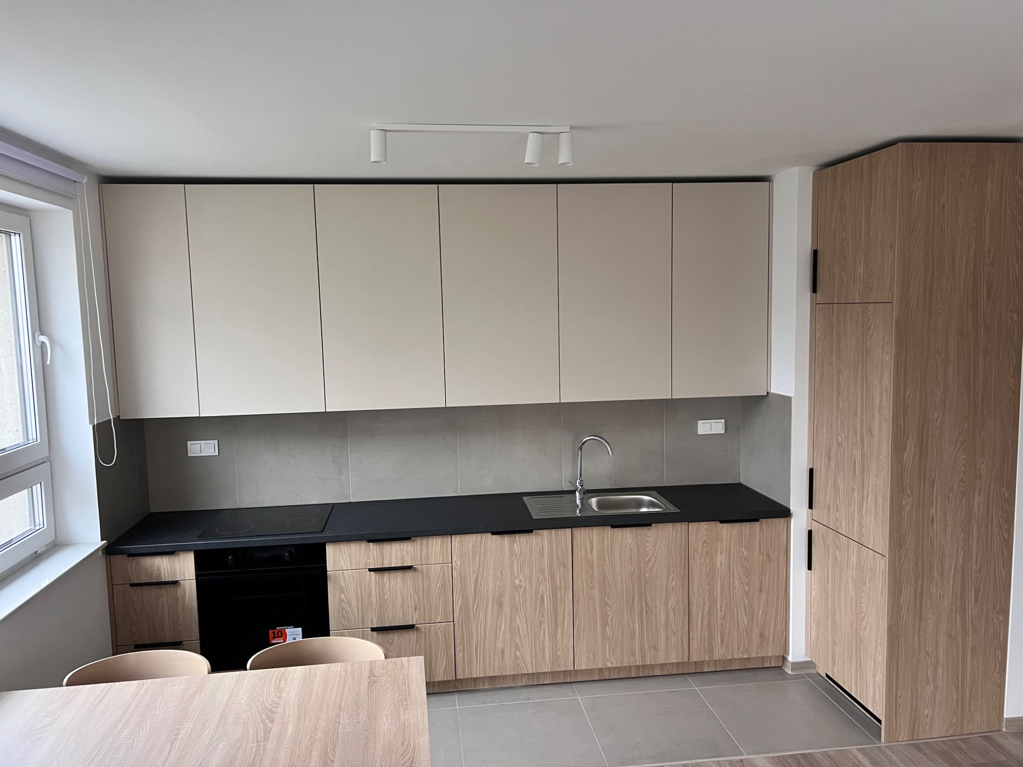 Jasna, nowoczesna kuchnia z drewnianymi szafkami i czarnym blatem. Widoczne okno, zlew i płyta grzewcza. Minimalistyczne oświetlenie sufitowe.