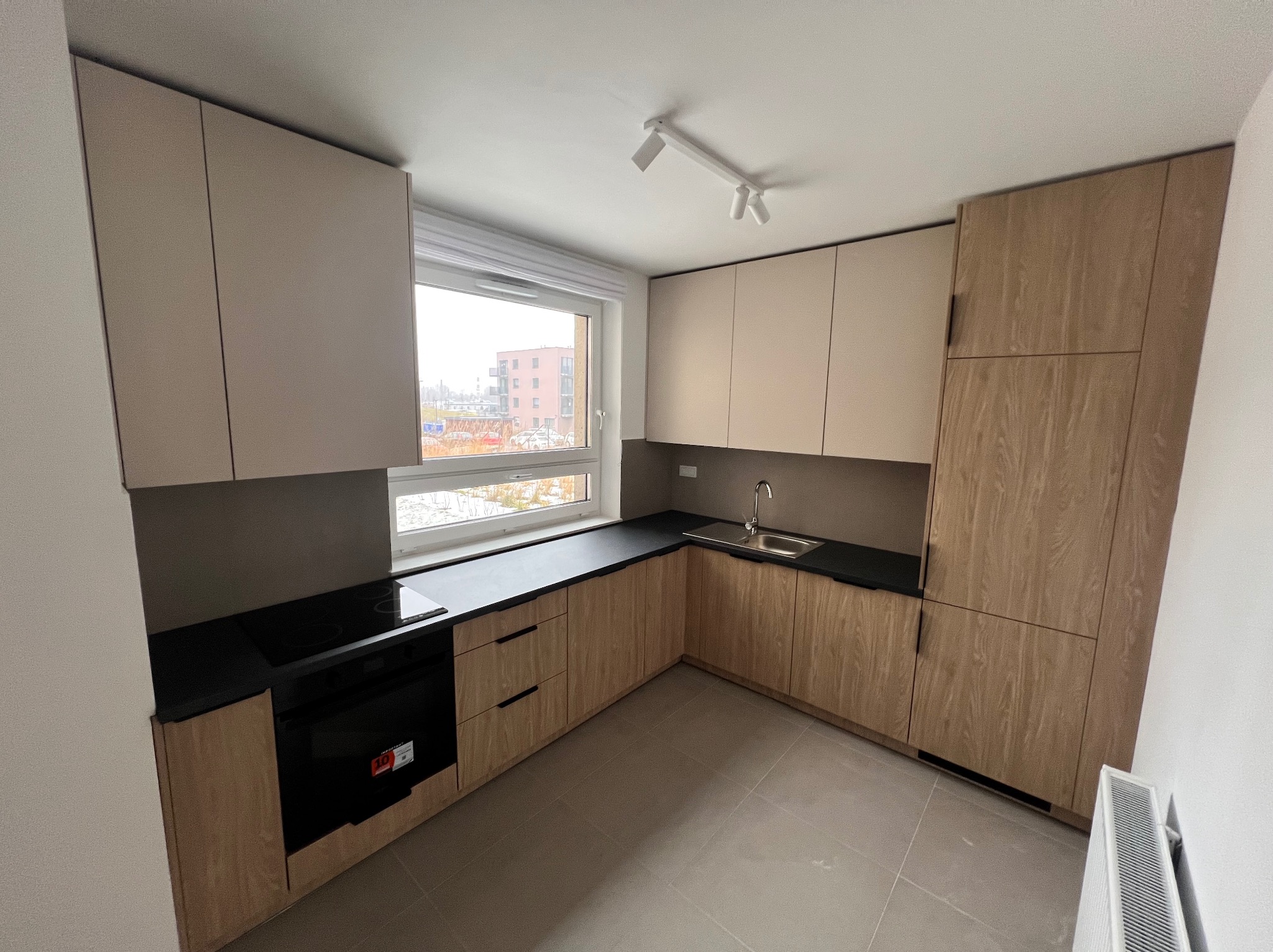 Jasna kuchnia z drewnianymi szafkami i czarnym blatem w Makowie Podhalańskim. Nowoczesne oświetlenie sufitowe i widok z okna na budynek w tle.