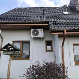 SO-FAR SPÓŁKA Z OGRANICZONĄ ODPOWIEDZIALNOŚCIĄ - Biały budynek z ciemną blachodachówką, brązowymi rynnami i zamontowaną jednostką zewnętrzną klimatyzacji LG na ścianie, widok z dołu na tle błękitnego nieba, karmnik dla ptaków w kadrze.