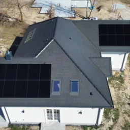 Widok z góry na dom z grafitowym dachem pokrytym panelami fotowoltaicznymi. Na dachu okna połaciowe. W tle drzewa i teren budowy.