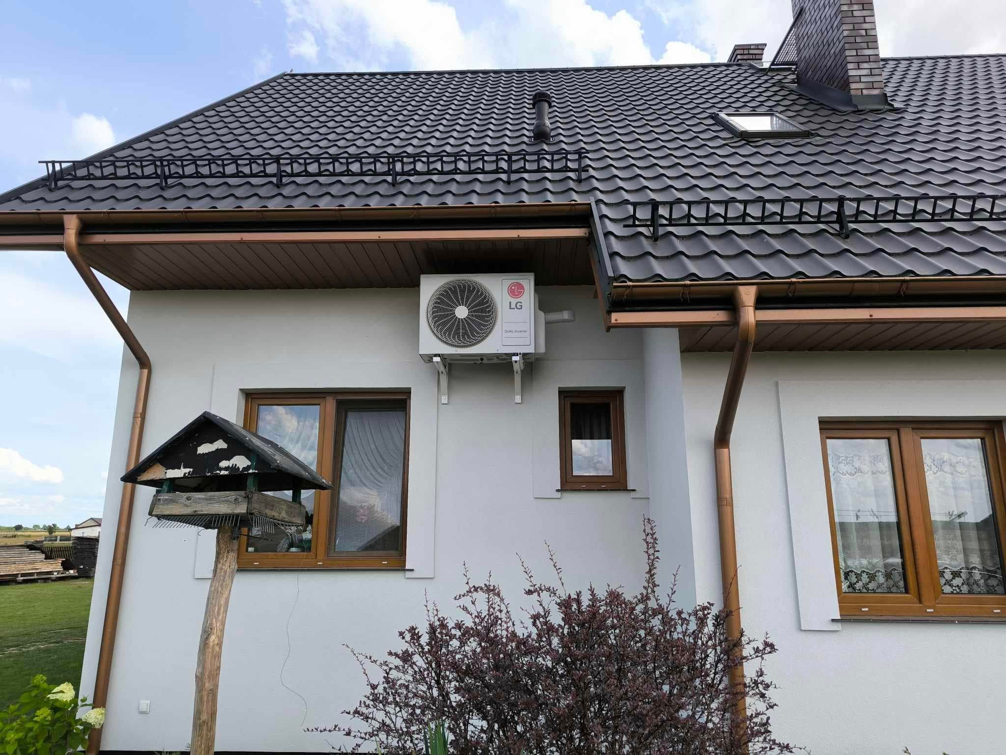 Biały budynek z ciemną blachodachówką, brązowymi rynnami i zamontowaną jednostką zewnętrzną klimatyzacji LG na ścianie, widok z dołu na tle błękitnego nieba, karmnik dla ptaków w kadrze.
