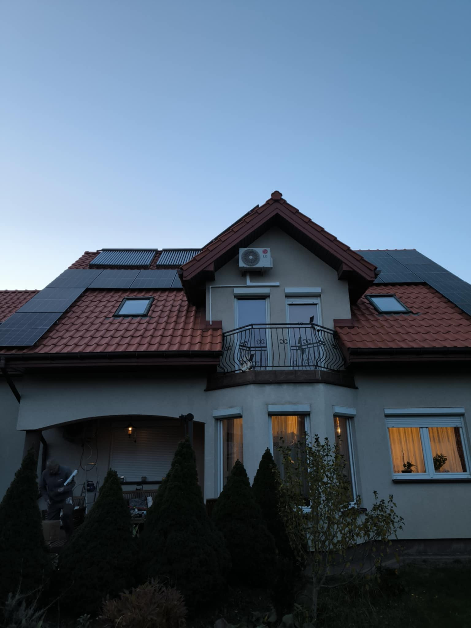 Dom jednorodzinny z panelami fotowoltaicznymi na dachu i kolektorami słonecznymi, klimatyzator LG na elewacji, widok z dołu na tle wieczornego nieba.