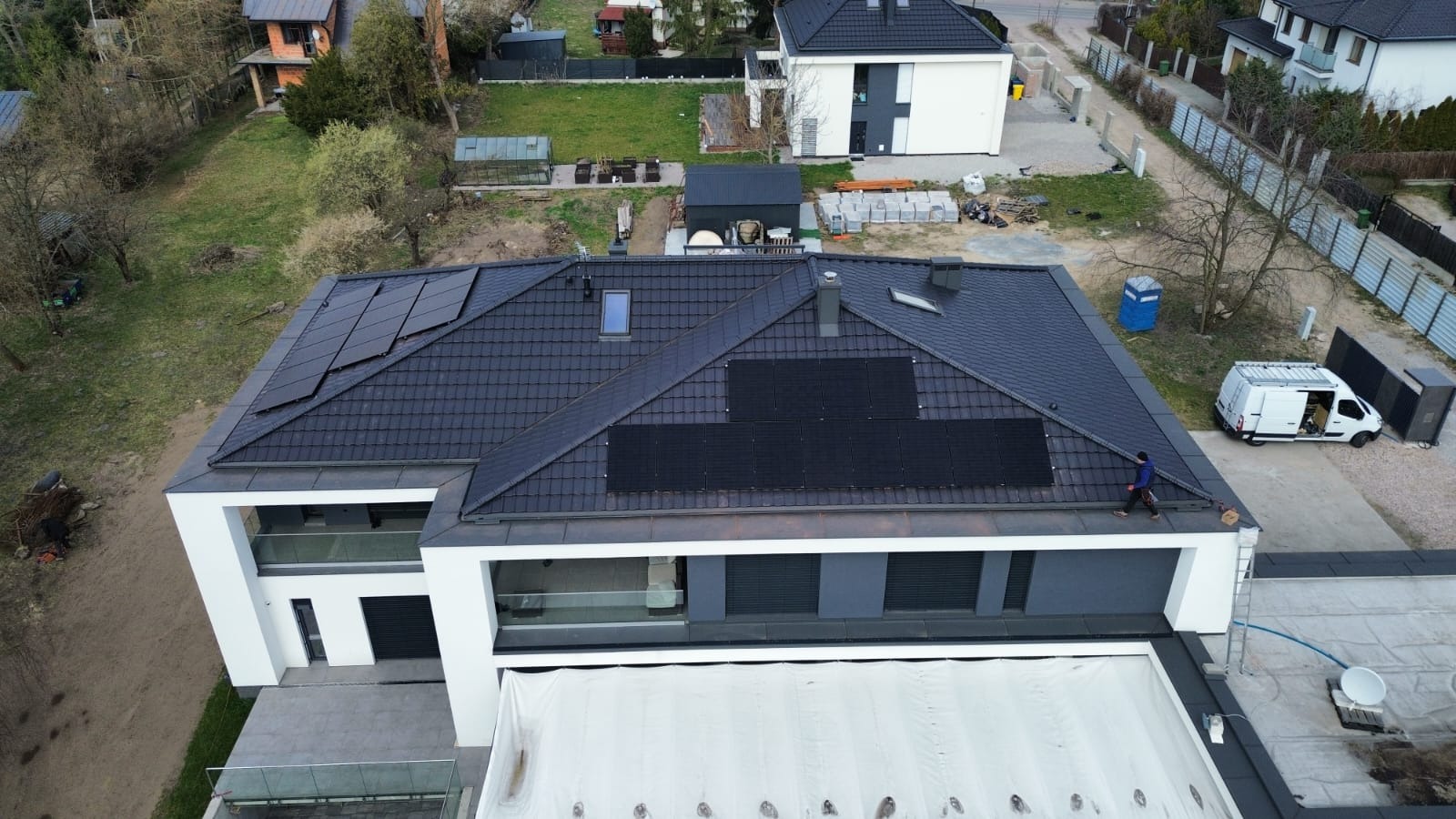 Montaż paneli słonecznych na dachu nowoczesnego domu z ciemną dachówką. Widoczny pracownik w trakcie instalacji, biały van i otoczenie budynku.