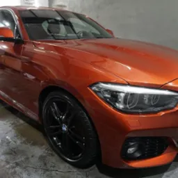Błyszczący, pomarańczowy BMW serii 1 z czarnymi felgami, po umyciu w myjni, widoczne krople wody na karoserii i mokra posadzka.