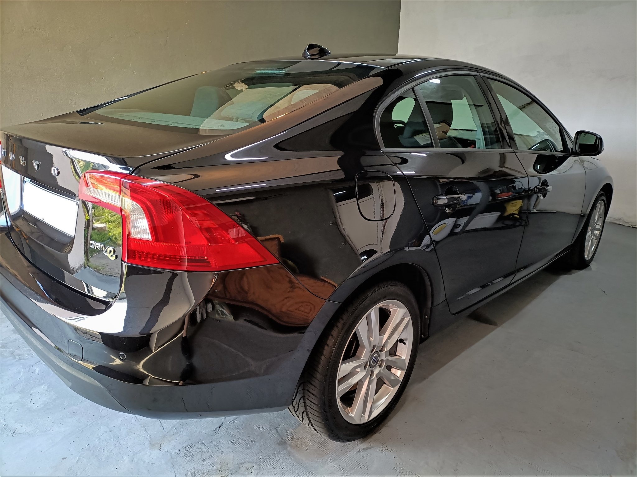 Błyszczący, czarny samochód Volvo S60 po umyciu, widoczny z tyłu pod kątem. Na lakierze widoczne odbicia otoczenia. W tle szara ściana garażu.