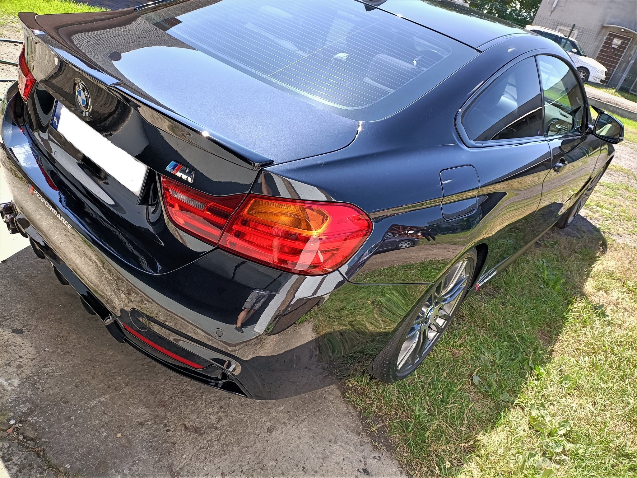 Błyszczący, czarny samochód BMW M4 z widocznym logo M Performance, zaparkowany na granicy betonu i trawy, po umyciu.