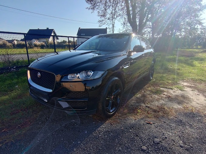 Błyszczący, czarny Jaguar F-Pace zaparkowany na żwirowej nawierzchni w słoneczny dzień, widoczny z przodu, z odbiciami w karoserii i elementami krajobrazu w tle.