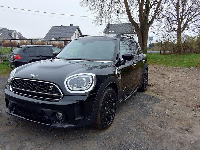 Czarny Mini Countryman z napędem hybrydowym zaparkowany na żwirowym podłożu, widoczny z przodu pod kątem, z domami w tle