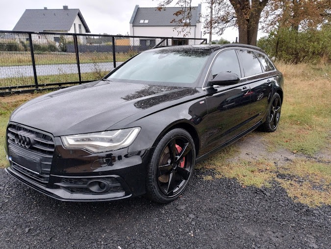 Czarny samochód Audi A6 Avant po umyciu, z widocznymi czerwonymi zaciskami hamulcowymi i kroplami wody na lakierze, zaparkowany na żwirowym podłożu przed domem.