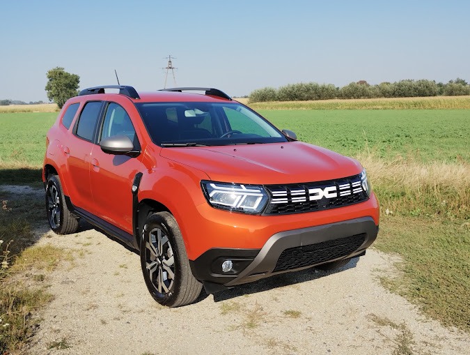 Pomarańczowy samochód typu SUV, Dacia Duster, zaparkowany na żwirowej drodze z polami w tle, widok z przodu.