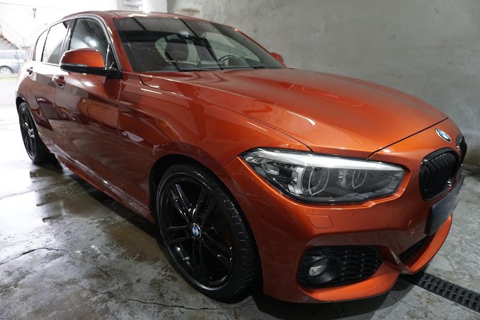 Błyszczący, pomarańczowy BMW serii 1 z czarnymi felgami, po umyciu w myjni, widoczne krople wody na karoserii i mokra posadzka.