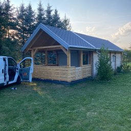 Piotr Syczewski - Nowoczesna, drewniana konstrukcja z szarym dachem, prawdopodobnie altana lub domek letniskowy, z częściowo otwartymi ścianami i drewnianą balustradą, obok zaparkowany biały samochód dostawczy...