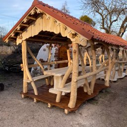 Piotr Syczewski - Rustykalna konstrukcja z drewnianych bali z czerwoną dachówką, tworząca zadaszone miejsce do siedzenia z drewnianym stołem i ławkami, na tle ogrodu z piecem chlebowym.