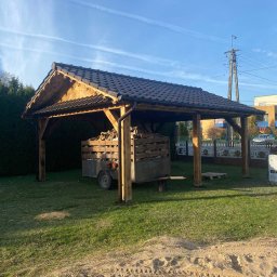 Piotr Syczewski - Zadaszona konstrukcja z drewna z ciemną dachówką, pod którą stoi przyczepa z drewnem opałowym, widok z trawnika.