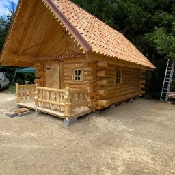 Piotr Syczewski - Drewniana konstrukcja z bali, prawdopodobnie domek letniskowy lub mała sauna, z ozdobnym dachem i małym tarasem z balustradą, umieszczona na betonowych fundamentach. W tle widoczne drzewa i drabina.