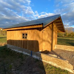 Piotr Syczewski - Drewniana konstrukcja z metalowym dachem i małym oknem, w tle łąka i drzewa, oświetlona popołudniowym słońcem.