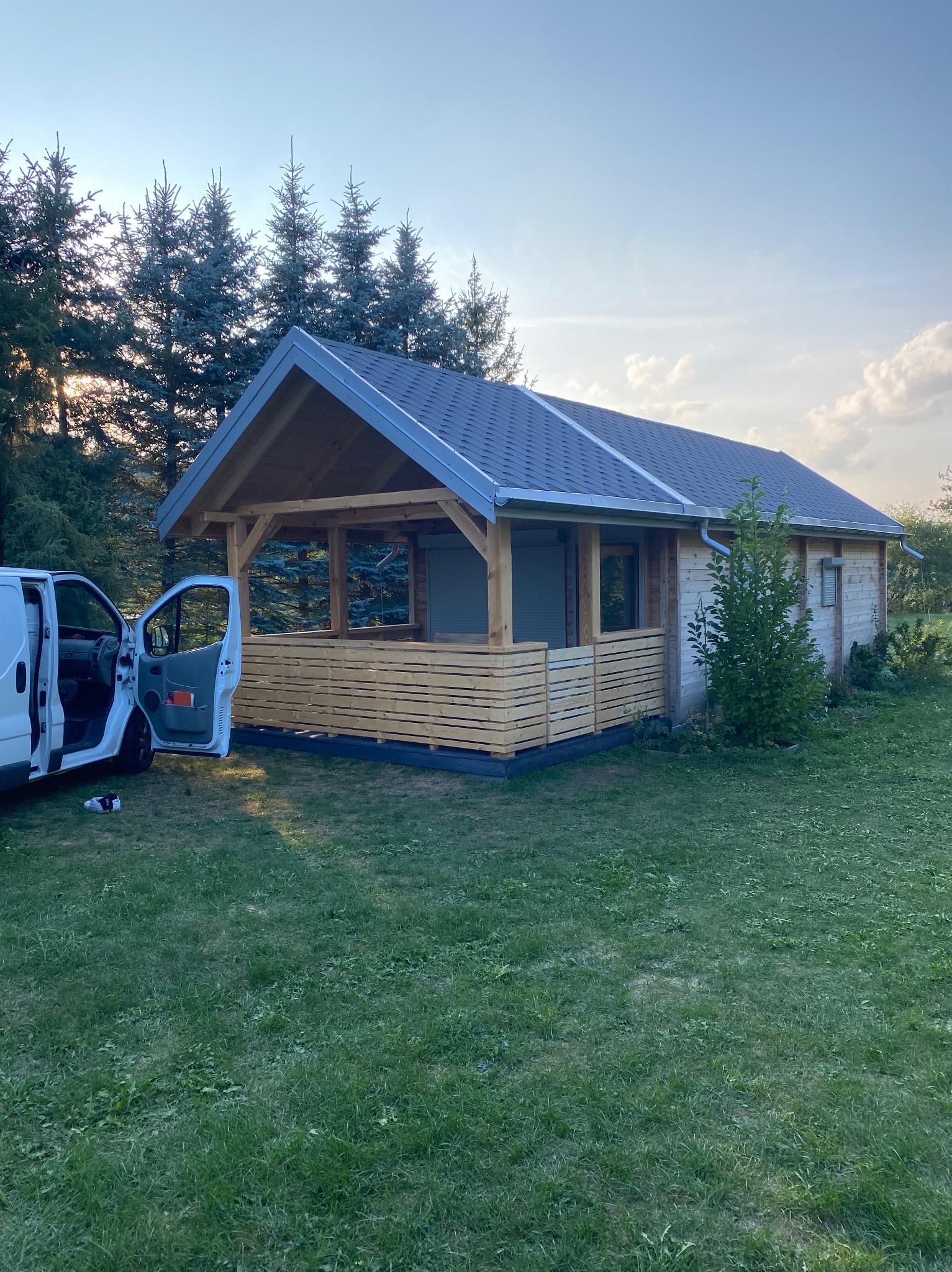 Nowoczesna, drewniana konstrukcja z szarym dachem, prawdopodobnie altana lub domek letniskowy, z częściowo otwartymi ścianami i drewnianą balustradą, obok zaparkowany biały samochód dostawczy...