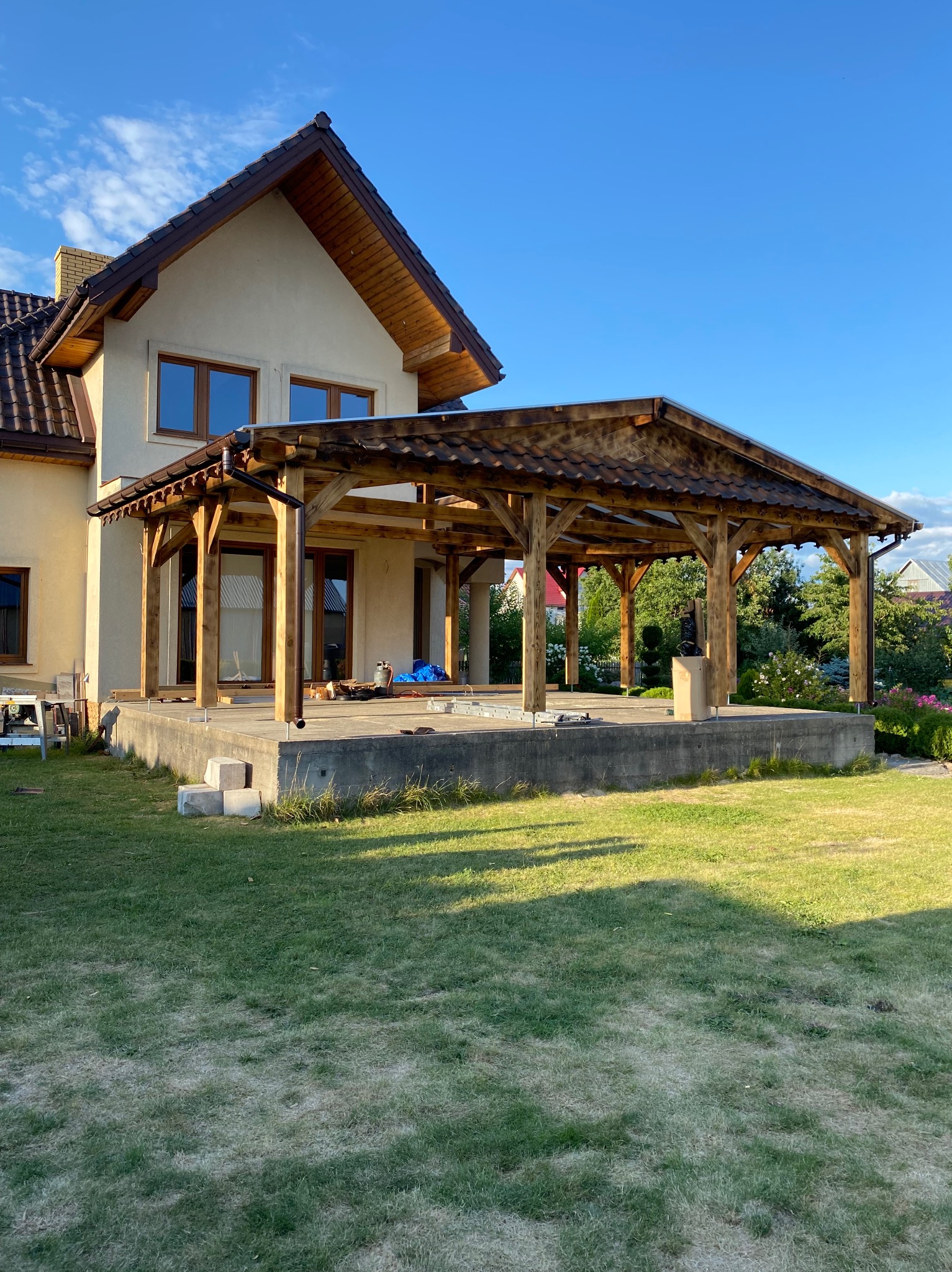 Drewniana konstrukcja z zadaszeniem, prawdopodobnie w trakcie budowy, przylegająca do domu z widocznymi narzędziami i materiałami budowlanymi na betonowej płycie.