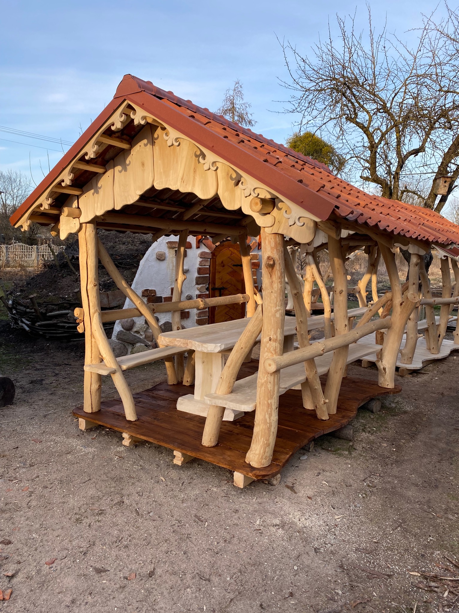 Rustykalna konstrukcja z drewnianych bali z czerwoną dachówką, tworząca zadaszone miejsce do siedzenia z drewnianym stołem i ławkami, na tle ogrodu z piecem chlebowym.