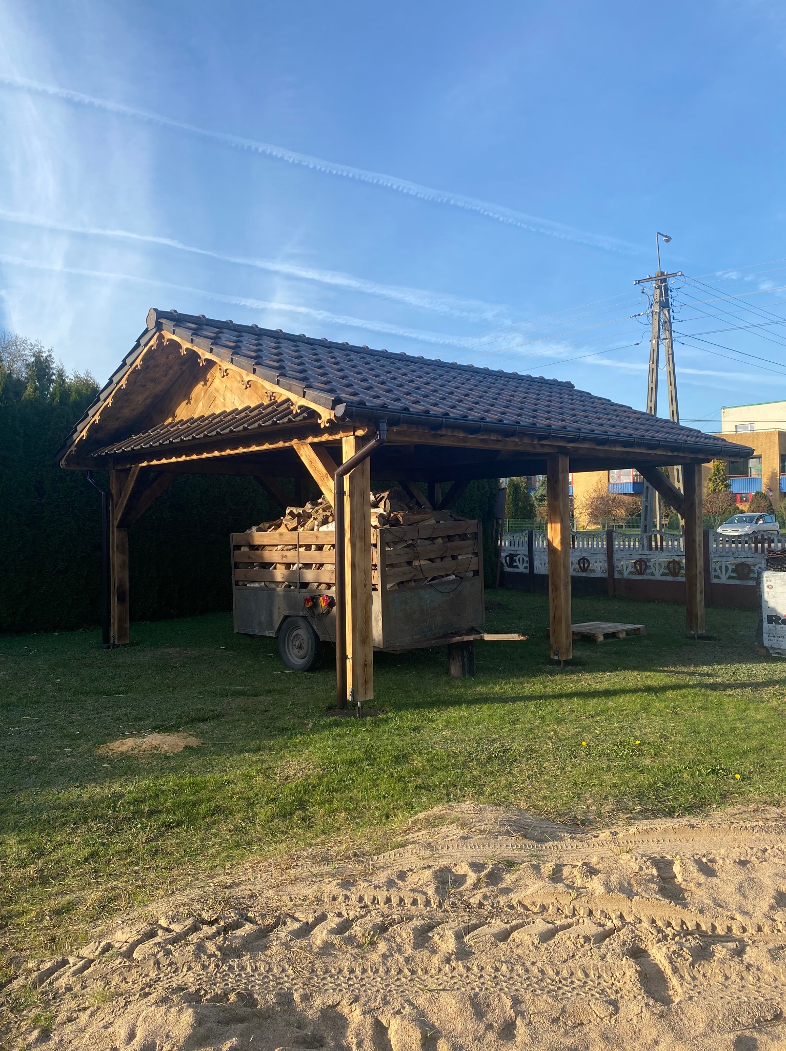 Zadaszona konstrukcja z drewna z ciemną dachówką, pod którą stoi przyczepa z drewnem opałowym, widok z trawnika.